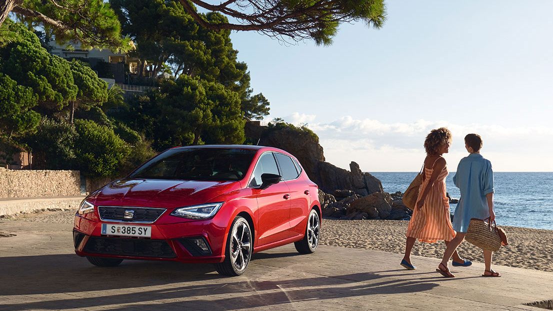 Ein roter SEAT Ibiza steht neben einem Strand und zwei Freundinnen gehen gerade Richtung Meer 
