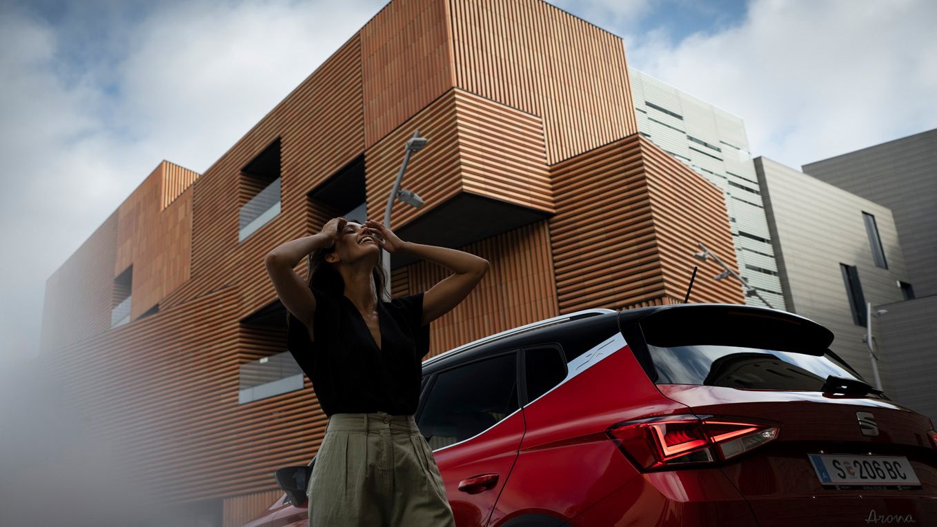 Eine Frau steht vor einem roten SEAT Arona mit schwarzem Dach, der aus der Heckperspektive zu sehen ist