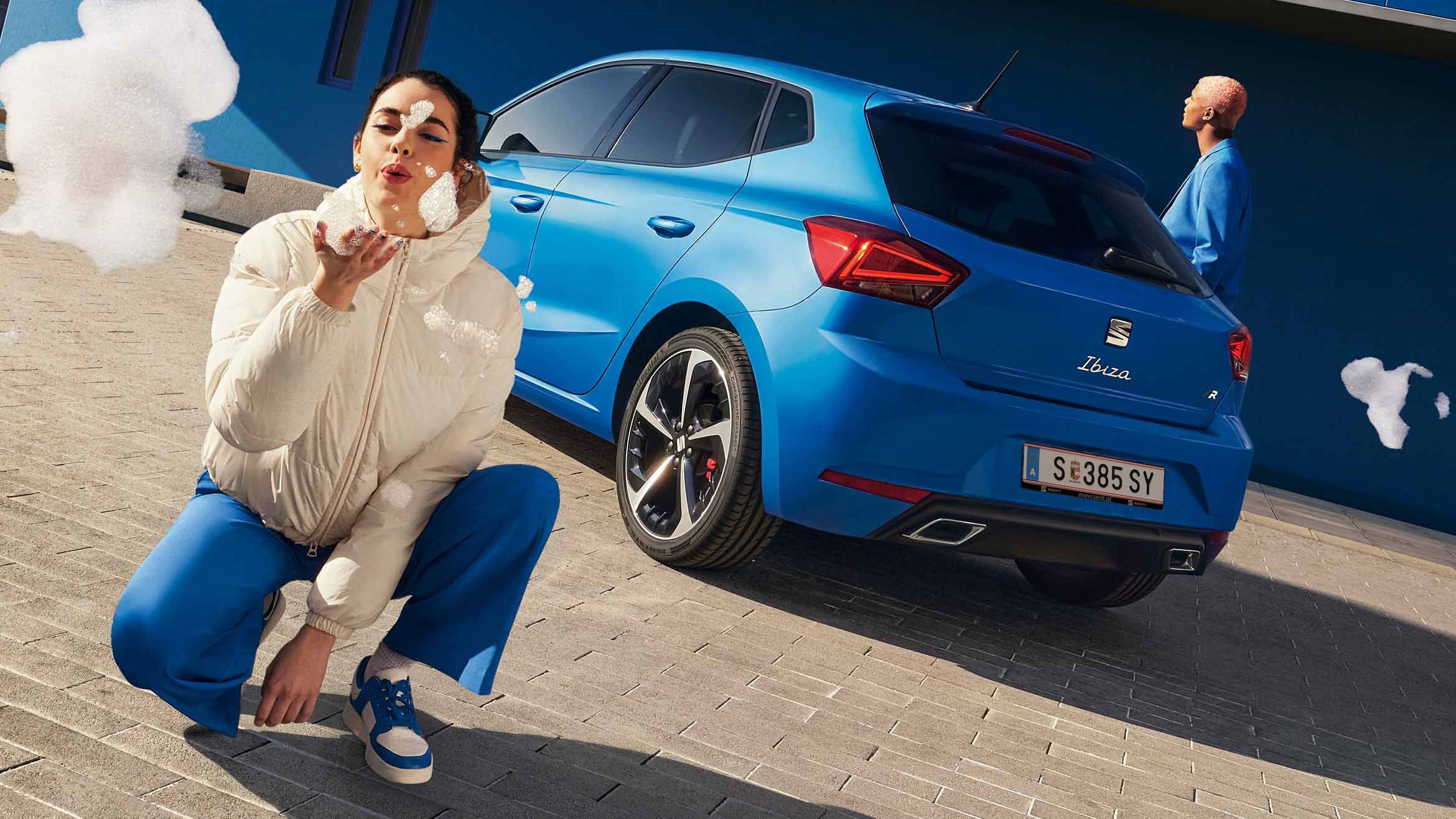 Ein blauer SEAT Ibiza steht mit dem Heck zu einem blauen Gebäude. Ein Mann im blauen Anzug geht daran vorbei, während eine Frau direkt vor der Kamera kniet und Seifenblasen in Richtung Linse pustet.