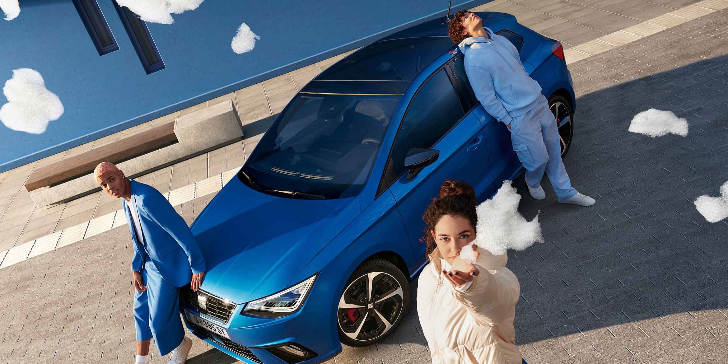 Ein blauer SEAT Ibiza aus der Vogelperspektive: Drei Personen, alle in blauer Kleidung, posieren am Auto – zwei lehnen an der Motorhaube, eine an der Seitentür. Eine der Personen hält Schaum in die Kamera.