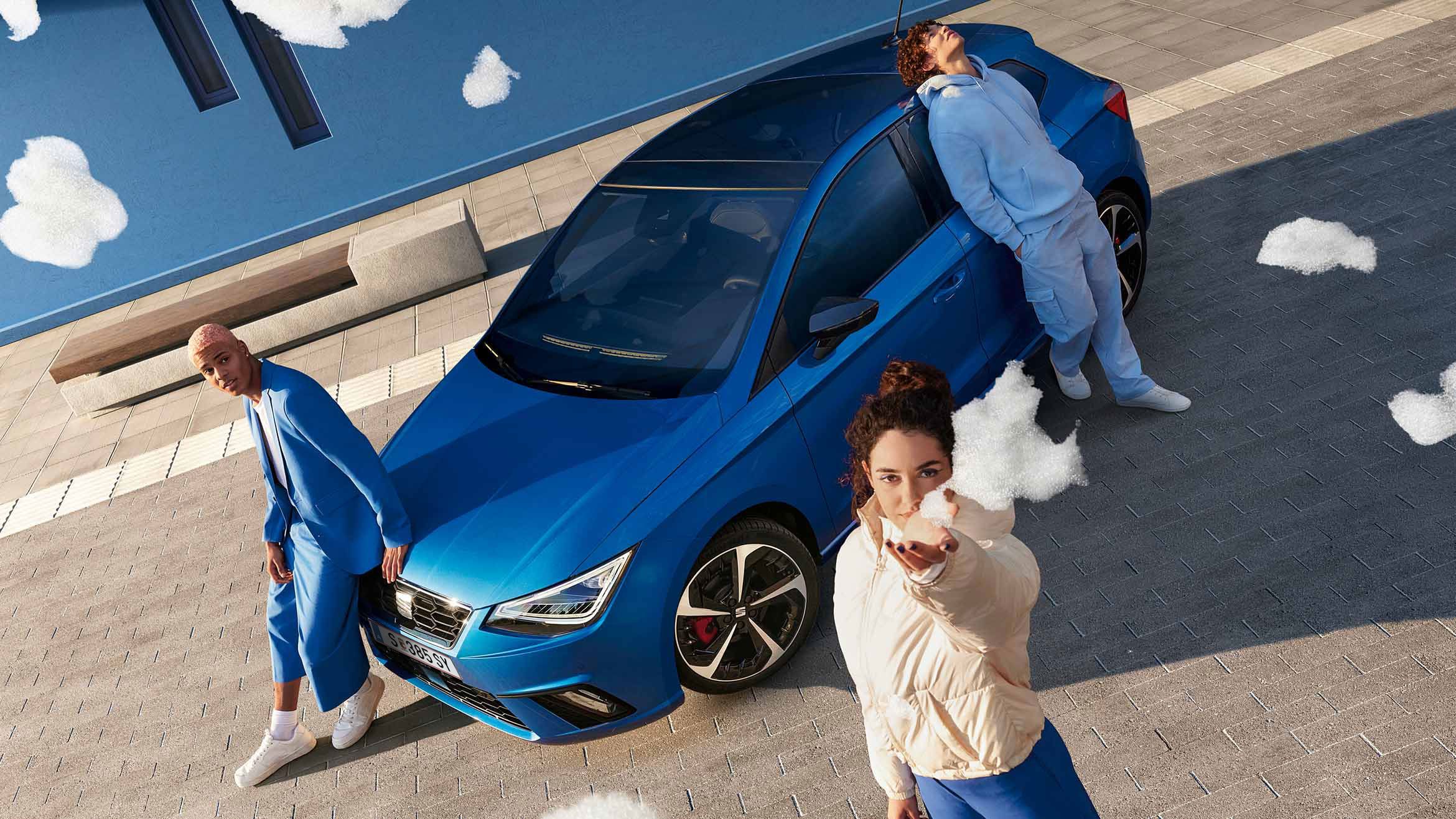 Ein blauer SEAT Ibiza aus der Vogelperspektive: Drei Personen, alle in blauer Kleidung, posieren am Auto – zwei lehnen an der Motorhaube, eine an der Seitentür. Eine der Personen hält Schaum in die Kamera.
